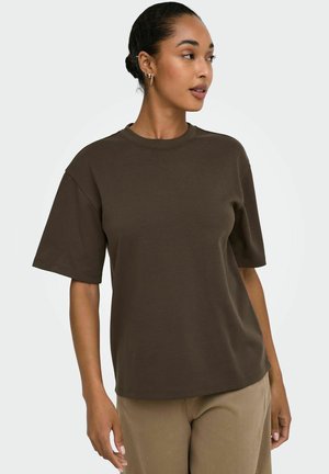 Femme aux cheveux bruns en chignon, portant des boucles d'oreilles anneaux dorées, un t-shirt marron foncé à manches courtes et un pantalon marron clair, regardant vers sa droite.