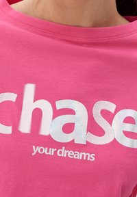 Rosa bomullströja med stor vit och iriserande text "jaga dina drömmar" med rund halsringning och korta ärmar.