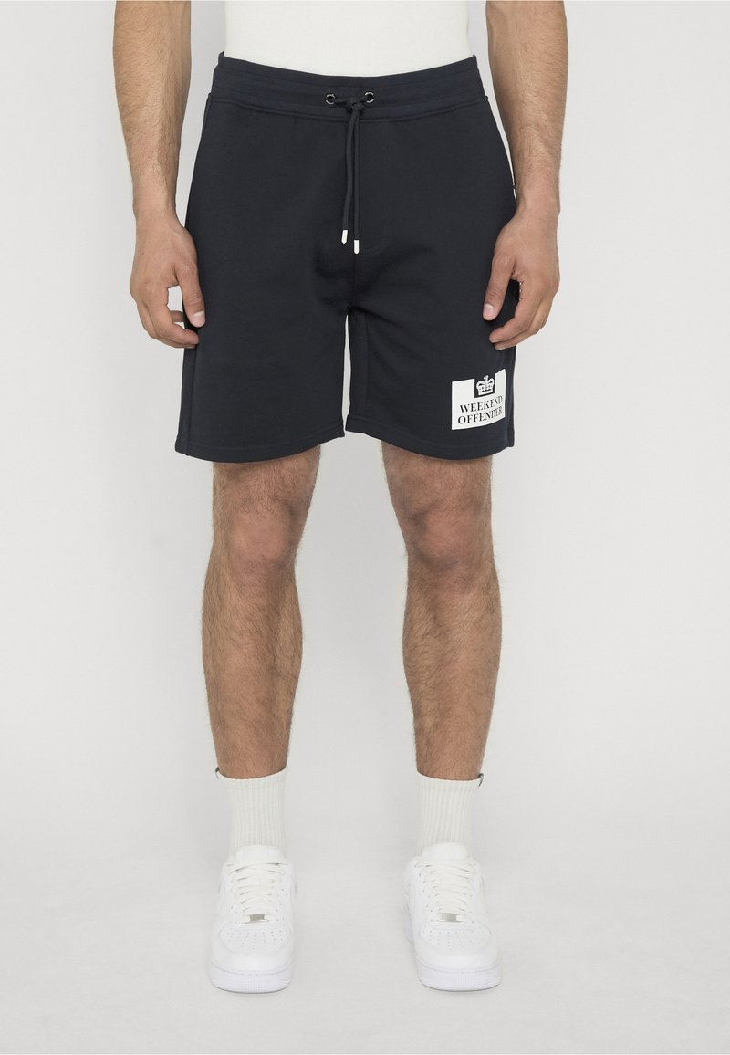 Μαύρα σορτς με κορδόνι και λευκό λογότυπο "Weekend Offender", φορεμένα από ένα άτομο που στέκεται με λευκές κάλτσες και λευκά αθλητικά παπούτσια.