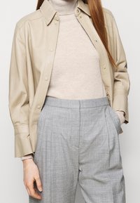Veste chemise en cuir beige avec un col et des boutons-pression, superposée à un pull col roulé gris clair et un pantalon à carreaux gris clair.