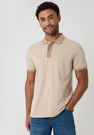 Uomo con polo a maniche corte beige e bianca a righe e jeans blu, in piedi con una mano in tasca su uno sfondo uniforme.