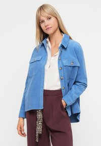 Γαλάζιο σακάκι από καμπαρντίνα με κουμπιά στο μπροστά μέρος, δύο τσέπες στο στήθος και χαλαρή γραμμή. Φορεμένο πάνω από ένα λευκό πουκάμισο και σκούρες μοβ παντελόνες.