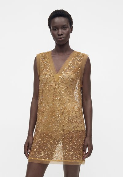 Femme portant une robe dorée sans manches, transparente, ornée de sequins et de perles complexes, se tenant devant un fond blanc uni.