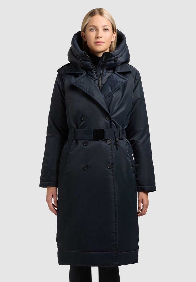 Temno modri dolg plašč z dvojnimi zapenjanji, velikim kapuco, pasom v predelu pasu in gladko, sijočo teksturo. Ima zadrgo in gumbno zapiranje.