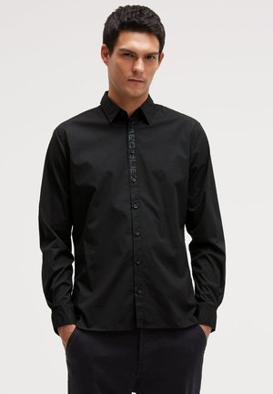 Mann trägt ein schwarzes, langärmliges Hemd mit subtilen vertikalen Textdetails, steht mit den Händen in den Taschen vor einem einfarbigen Hintergrund.