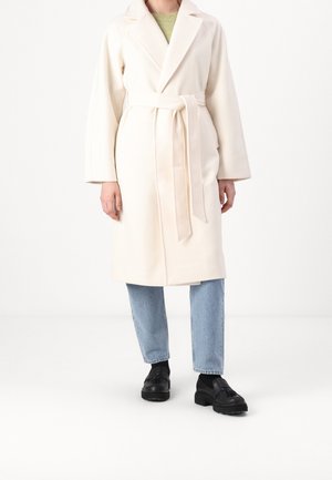 Cremefarvet robe med bælte i taljen, brede ærmer og krave med spring, parret med lys blå jeans og sorte sko.