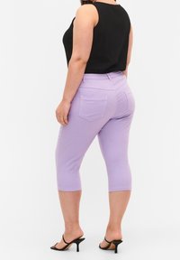 Pantaloni capri viola con tasche posteriori, texture liscia e design aderente, abbinati a un top nero senza maniche e scarpe aperte.