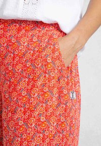 Red floral patterned trousers with a pocket. Design features orange, yellow, and blue flowers on a bright red background. Soft fabric texture.