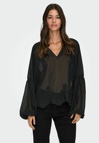 Blusa nera trasparente con motivo a pois, scollatura a v e maniche a palloncino voluminose. Presenta un design floreale ricamato all'orlo.