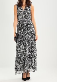 Robe maxi avec un décolleté en V, sans manches, en rayures zèbre noires et blanches, mettant en avant une taille cintrée et une jupe fluide. Talons noirs et pochette.