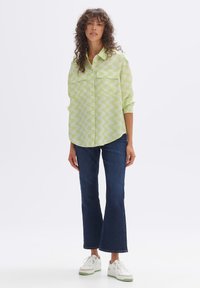 Green patterned button-up shirt with two chest pockets, paired with dark blue flared jeans and white sneakers with green accents.