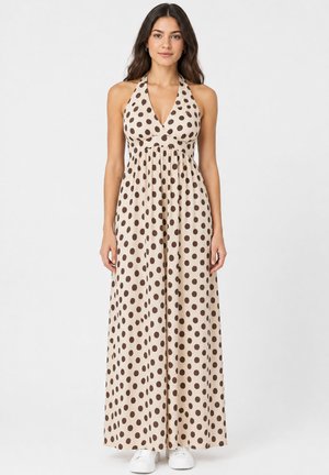 Vrouw draagt een beige halternek maxi jurk met grote donkerbruine polkadots en witte sneakers, staand tegen een effen achtergrond.