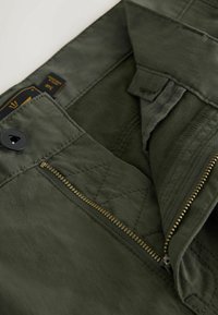 Dark green pants with an open brass zipper, black button, and visible size label inside showing "34R."