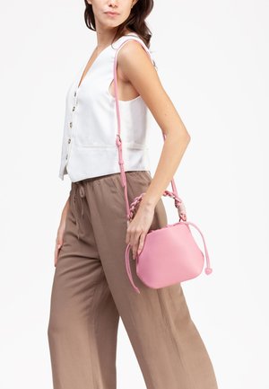 Donna che indossa un gilet bianco senza maniche abbottonato e pantaloni marroni a gamba larga, con una piccola borsa rosa con manico intrecciato e tracolla.