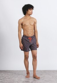 Bărbat muscular cu păr creț stă desculț, purtând șorturi de baie bleumarin cu un model floral și un șnur portocaliu, privind spre dreapta.