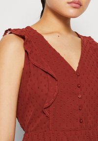 Zbliżenie na kobietę w bezrękawnikowej sukience w kolorze rdzawego czerwonego z teksturowymi kropkami, falbaniastymi detalami na ramionach oraz zapięciem w kształcie litery V z guzikami.
