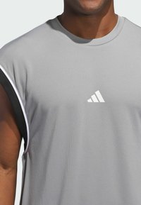 Camiseta deportiva gris con un acabado texturizado, que presenta un logo blanco de Adidas y un ribete negro contrastante a lo largo de las sisas y el cuello.