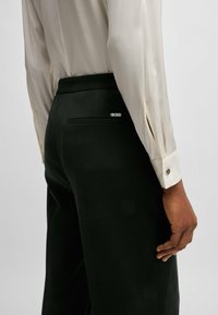 Pantaloni neri su misura con una texture liscia, dotati di una tasca posteriore e di un piccolo logo 'BOSS' bianco. Abbinati a una blusa color crema.