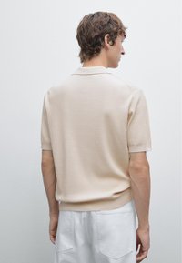 Beige, ribbet kortærmet polo skjorte med klassisk krave, med en tætsiddende kant og subtil teksturdetalje. Båret med hvide bukser.