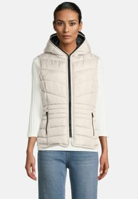 Vrouw in een lichtbeige gewatteerde hoodie-vest over een witte ribbelige lange mouwen top en blauwe spijkerbroek, recht vooruit kijkend.