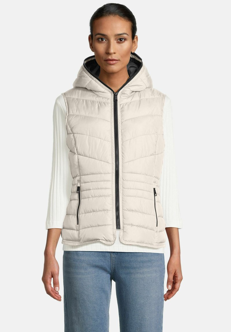 Vrouw in een lichtbeige gewatteerde hoodie-vest over een witte ribbelige lange mouwen top en blauwe spijkerbroek, recht vooruit kijkend.