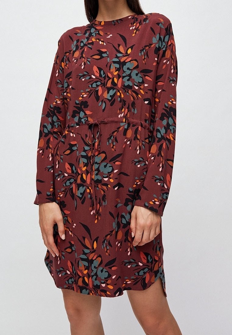 Femme portant une robe fleurie bordeaux à manches longues avec une taille à cordon, debout devant un fond clair uni.