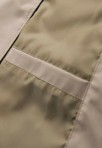 Nahaufnahme einer beigen Stofftasche mit einer genähten horizontalen Klappe auf einer glatten, leicht glänzenden Textiloberfläche.