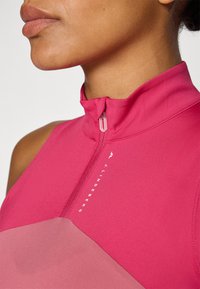 Top justo sem mangas em rosa brilhante com gola alta e fecho de zip, apresentando um painel em rosa mais claro em contraste e uma marcação vertical.