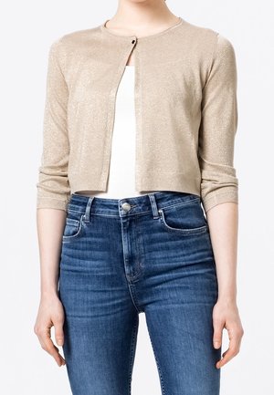 Kvinde iført en kort beige cardigan med trekvartærmer over en hvid top, kombineret med højtaljede blå jeans.