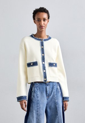 Cardigan corto bianco con rifiniture in denim, dotato di chiusura a bottoni e due tasche frontali. Il tessuto appare morbido con una finitura leggermente testurizzata.