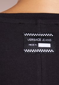 Étiquette en tissu noir affichant "VERSACE JEANS" avec des accents de couture blanche et un espace vierge pour les détails ; attachée à un vêtement noir texturé.