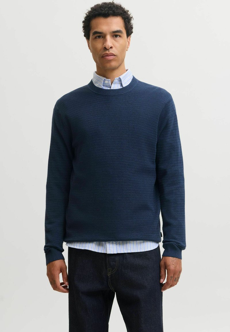 Homme portant un pull texturé bleu marine par-dessus une chemise à rayures bleu clair et blanc avec un jean foncé, debout devant un fond uni.