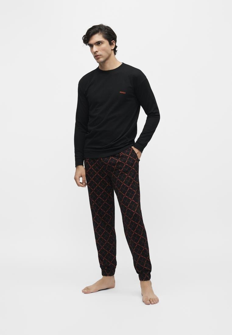 Homme portant une chemise noire à manches longues avec un petit logo rouge et un pantalon noir avec un motif losange rouge, debout pieds nus sur fond blanc.