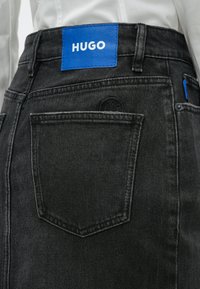 Jupe en denim noir avec une patch en cuir bleu portant l'étiquette "HUGO". Elle dispose de deux poches arrière et d'une couture distinctive le long de l'ourlet.