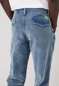 Homme portant un pantalon en velours côtelé bleu clair avec une poche arrière boutonnée et une chemise blanche rentrée, debout devant un fond uni.