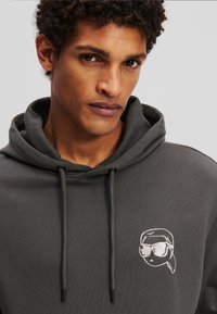 Hoodie gri realizat dintr-un material moale, având un guler cu șnur și o grafică albă brodată a unui personaj care poartă ochelari de soare pe piept.