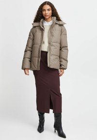 Puffjakke i taupe med høj krave og lynlås foran, parret med en beige striktrøje og mørk burgunder nederdel, suppleret med knæhøje sorte støvler.
