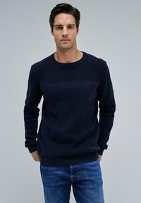 Navyblauer Strickpullover mit strukturiertem Mittelteil und gerippten Bündchen. Kombiniert mit blauen Jeans für einen lässigen Look.
