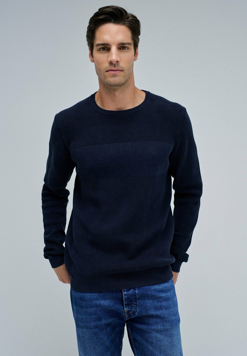 Navyblauer Strickpullover mit strukturiertem Mittelteil und gerippten Bündchen. Kombiniert mit blauen Jeans für einen lässigen Look.
