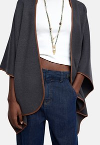 Szary, wełniany płaszcz z brązowym wykończeniem, w połączeniu z białą koszulką na ramiączkach i ciemnoniebieskimi dżinsami. Posiada warstwowy design i boczne kieszenie.