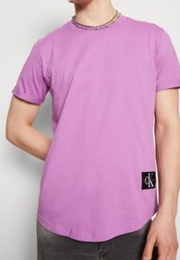 Osoba oblečená v lila tričku s krátkým rukávem s černou nášivkou loga Calvin Klein poblíž lemu a s stříbrným korálkovým náhrdelníkem.