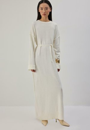 Femme portant une longue robe blanche en maille avec une ceinture, des manches retroussées, des escarpins beiges pointus et un gros bracelet en or à son poignet gauche.