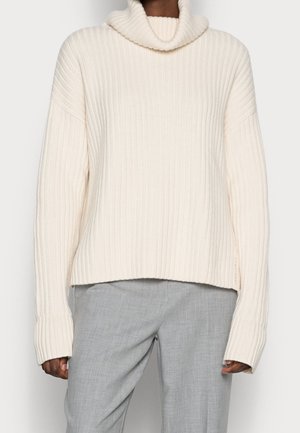 Personne portant un pull à col roulé côtelé de couleur crème avec des manches longues et un pantalon gris clair cintré.