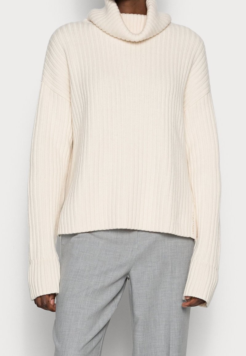 Personne portant un pull à col roulé côtelé de couleur crème avec des manches longues et un pantalon gris clair cintré.