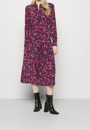 Kvinde iført knælængde langærmet kjole med pink og rød blomsterprint og sorte ankelstøvler med hæl, stående mod en ensfarvet baggrund.