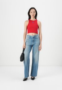 Rotes cropped Tanktop mit Logo, hellblaue weite Jeans, schwarze Handtasche und schwarze Schuhe. Ein schlichter Hintergrund hebt die Details des Outfits hervor.