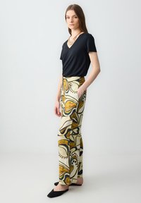 T-shirt noir à col en V associé à un pantalon crème avec des motifs abstraits audacieux en marron et bleu, et des chaussures noires à enfiler.