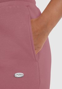 Pantalones jogger de color rosa claro con una textura suave, que cuentan con bolsillos laterales y un acento del logo plateado "Smilatex" cerca del bolsillo.