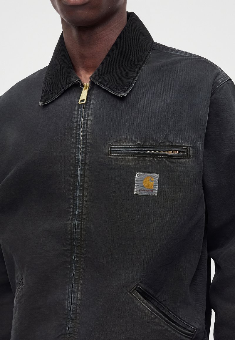 Nahaufnahme einer Person, die eine schwarze Carhartt-Jacke mit durchgehendem Reißverschluss, einer Reißverschlusstasche auf der Brust und einem sichtbaren Logo-Patch trägt.