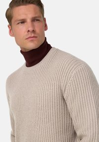 Pull en tricot beige avec un motif texturé et un col rond, superposé à un haut à col montant bordeaux foncé.